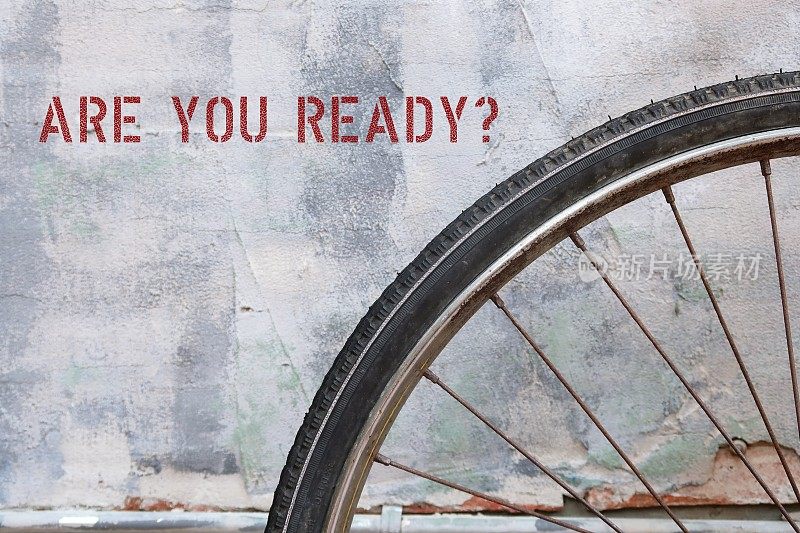 自行车车轮在水泥墙的背景上写着ARE YOU READY?用这个问题来判断某人是否准备做某事——开始紧张的活动或听到令人兴奋、震惊或惊讶的消息